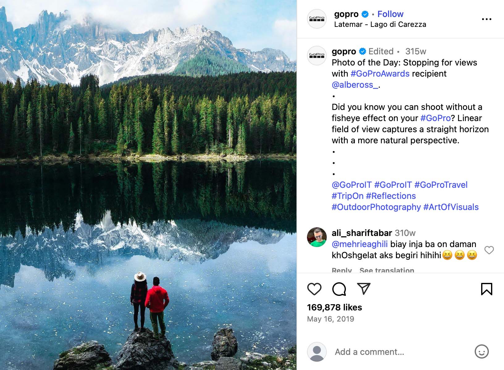 Two people standing by Lago di Carezza with forest and snowy mountains reflected in the water